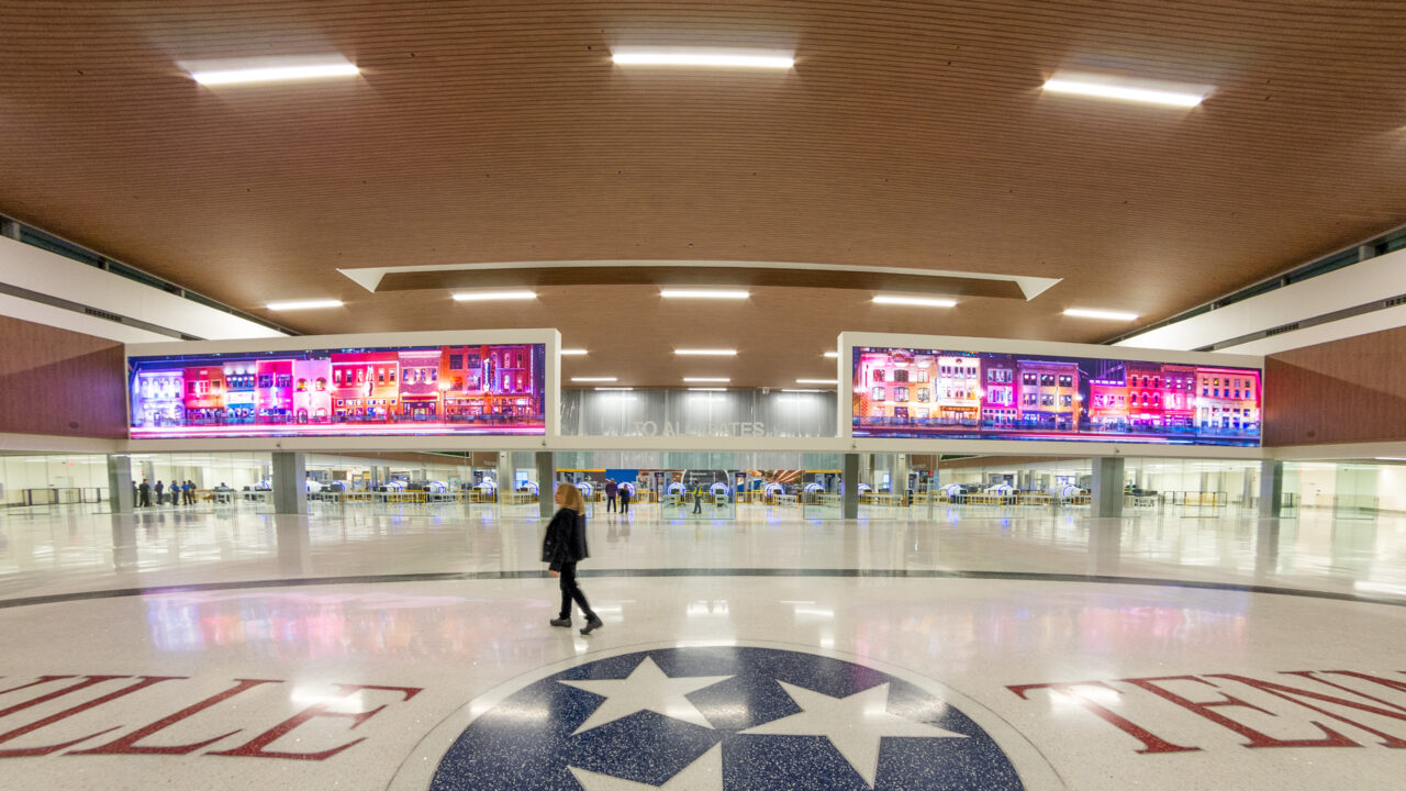 A woman is walking in the band new hall of the Nashville International Airport, with gigantic LED screens in the back.
